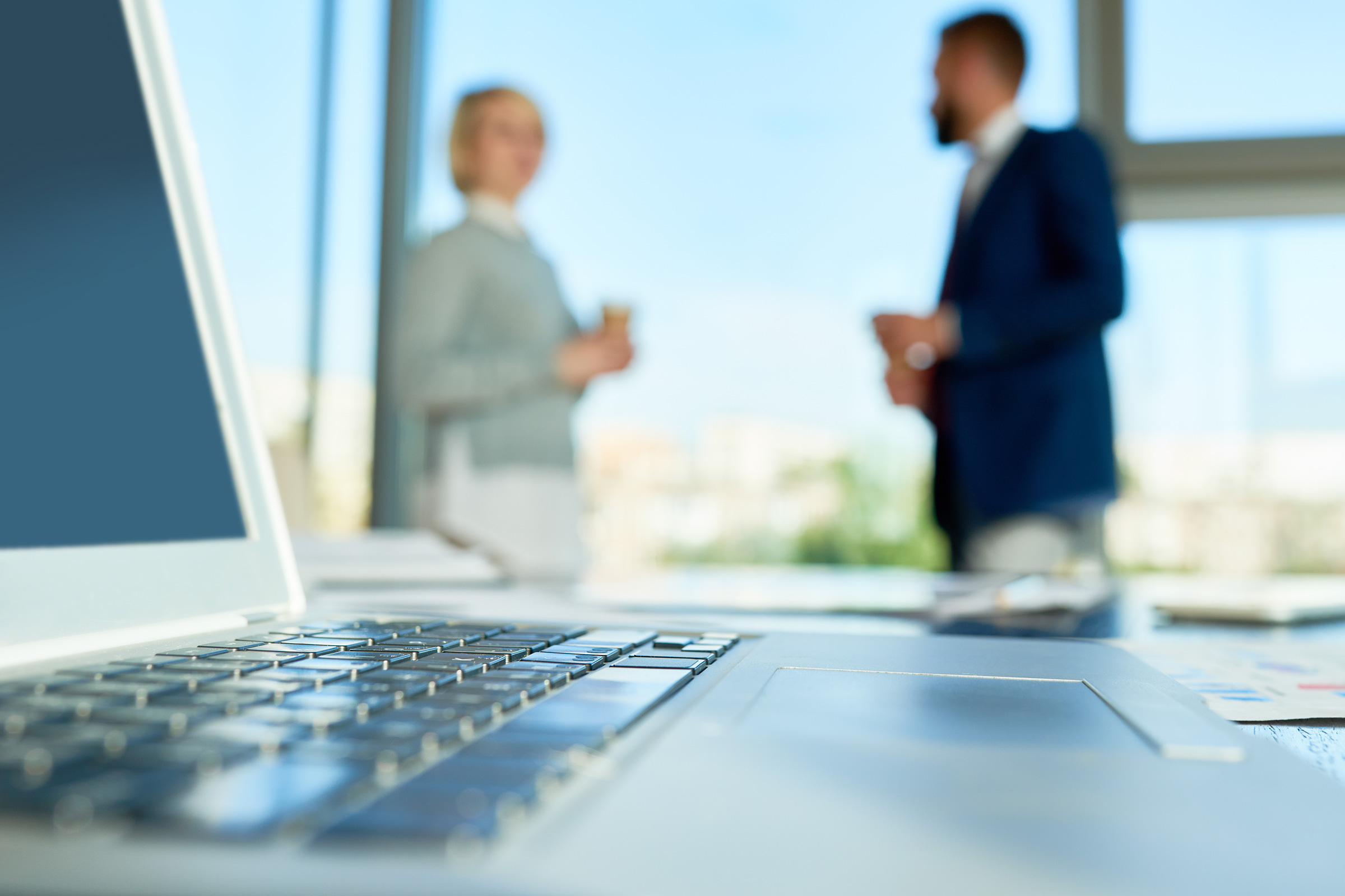 Laptop na biurku w nowoczesnym biurze podczas rozmowy rekrutacyjnej IT – dwoje profesjonalistów rozmawiających w tle przy panoramicznym oknie